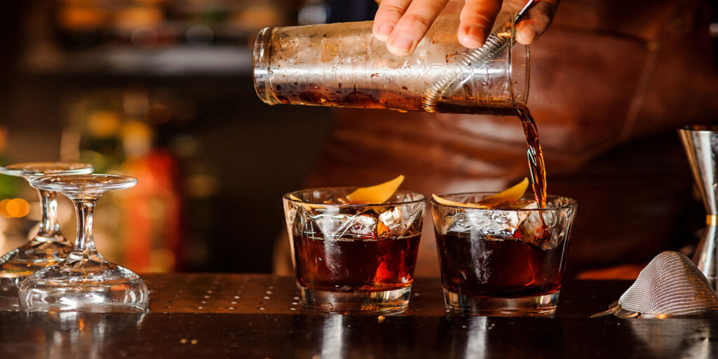 Bartender pouring fresh alcoholic drink into the glasses with ice cubes on the bar counter