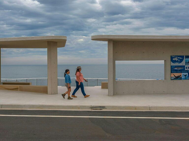 Two women walking towards whale information posters at Cape Solander lookout with ocean views.