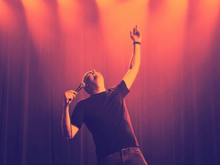 Tom holds a microphone to his mouth. His left hand is pointed upward and his gaze follows. He is wea