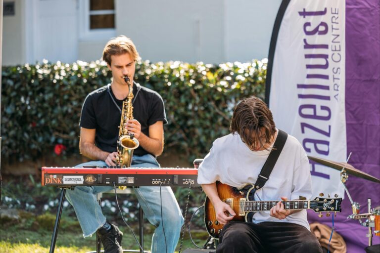 Two young musicians perform outdoors near a Hazelhurst Arts Centre banner.