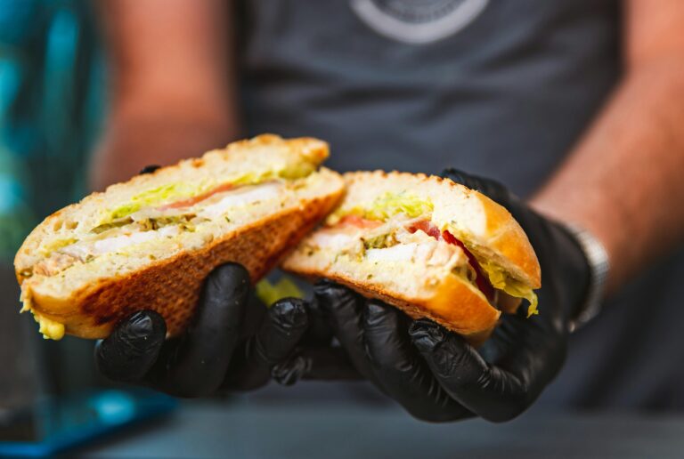 A person in black gloves holds out a filled sandwich cut in half, showing a toasted sandwich.