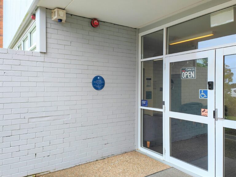Blue plaque on white painted wall, next to glass entrance doors of Denlea Cenrte building